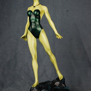 May include: A green-skinned figurine of a woman wearing a dark green swimsuit. The figurine is standing on a black rock base.