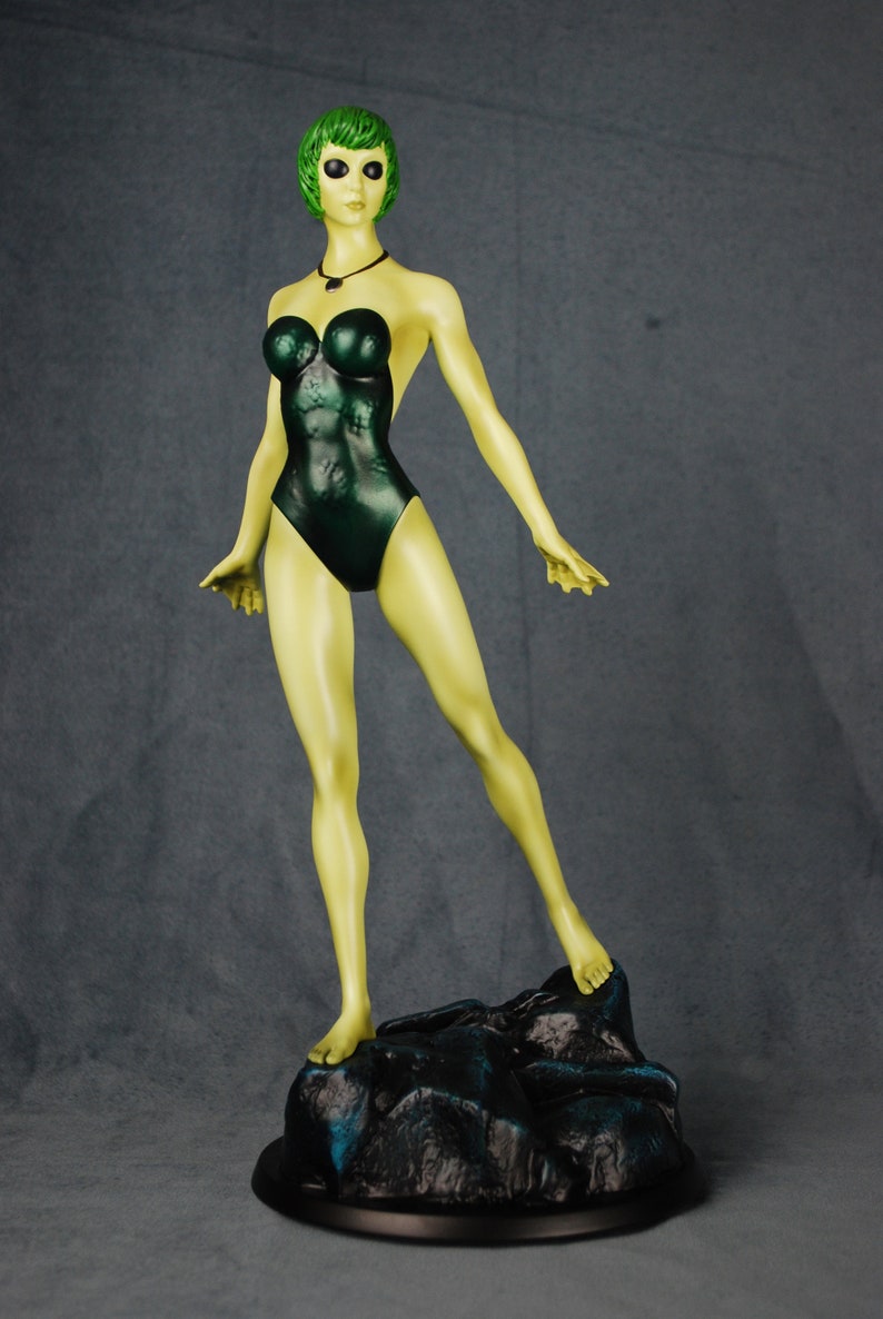 May include: A green-skinned figurine of a woman wearing a dark green swimsuit. The figurine is standing on a black rock base.