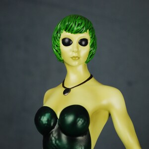 May include: A green-haired figurine with large black eyes, wearing a dark green strapless dress and a silver necklace with a pendant.