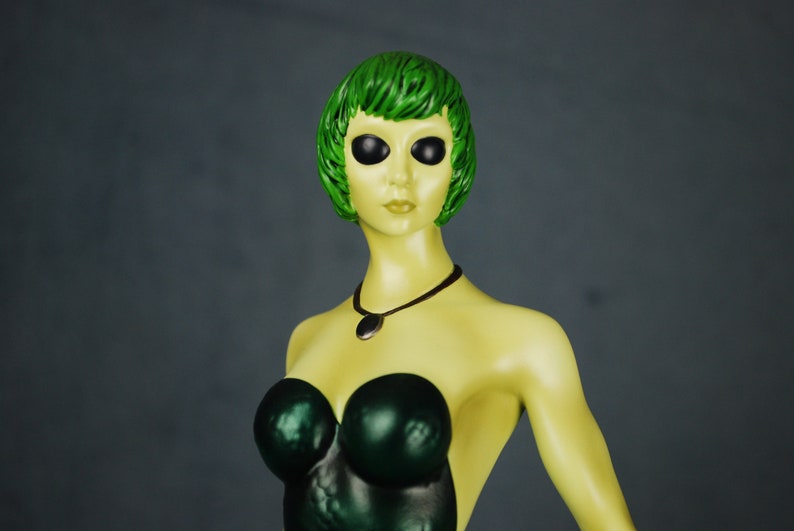 May include: A green-haired figurine with large black eyes, wearing a dark green strapless dress and a silver necklace with a pendant.