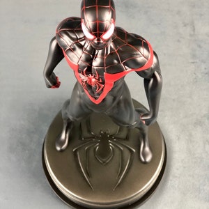May include: Black and red Spider-Man action figure with a black spider symbol on the base. The figure is standing with its arms raised and its head tilted down.