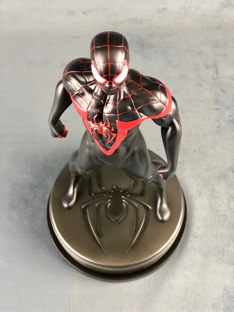 May include: Black and red Spider-Man action figure with a black spider symbol on the base. The figure is standing with its arms raised and its head tilted down.