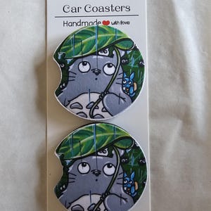 May include: Two car coasters featuring a gray cartoon character under a green leaf umbrella, with a smaller blue character. The coasters are on a white card with the text "Car Coasters Handmade with love."