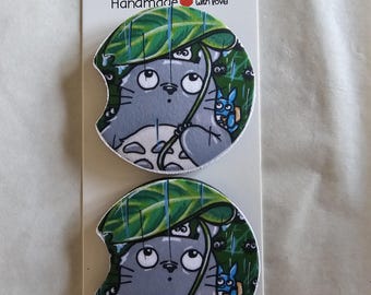 May include: Two car coasters featuring a gray cartoon character under a green leaf umbrella, with a smaller blue character. The coasters are on a white card with the text "Car Coasters Handmade with love."