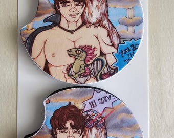 May include: Two round car coasters featuring a cartoon illustration of a shirtless man with brown hair, a small creature with pink hair, and a furry companion wearing glasses. The text reads "HI ZEV" and "You Be Us To". The coasters are on a white display card labelled "Car Coasters" and "Handmade with love".