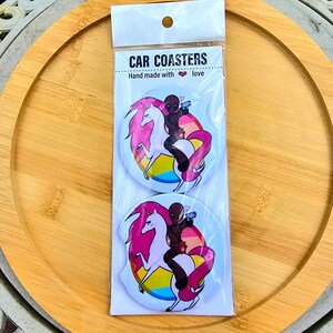 May include: Two round car coasters featuring a black figure riding a pink unicorn with a rainbow background. The text "CAR COASTERS" and "Hand made with love" are printed on the packaging.