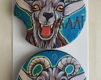 May include: Two circular car coasters featuring a graphic illustration of a screaming goat with orange eyes, grey fur, and curved horns against a blue background. The text AAAH is printed on the right side. The coasters are displayed on a white card labelled Car Coasters Handmade with love.