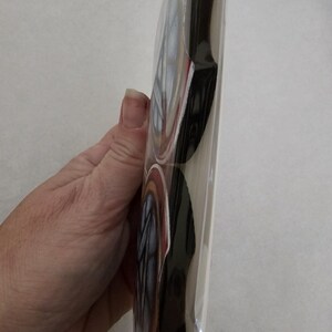 May include: A stack of round, decorative coasters in a clear plastic sleeve. The coasters have a design with red, white, and blue accents, and a black border. The coasters are held by a person's hand.
