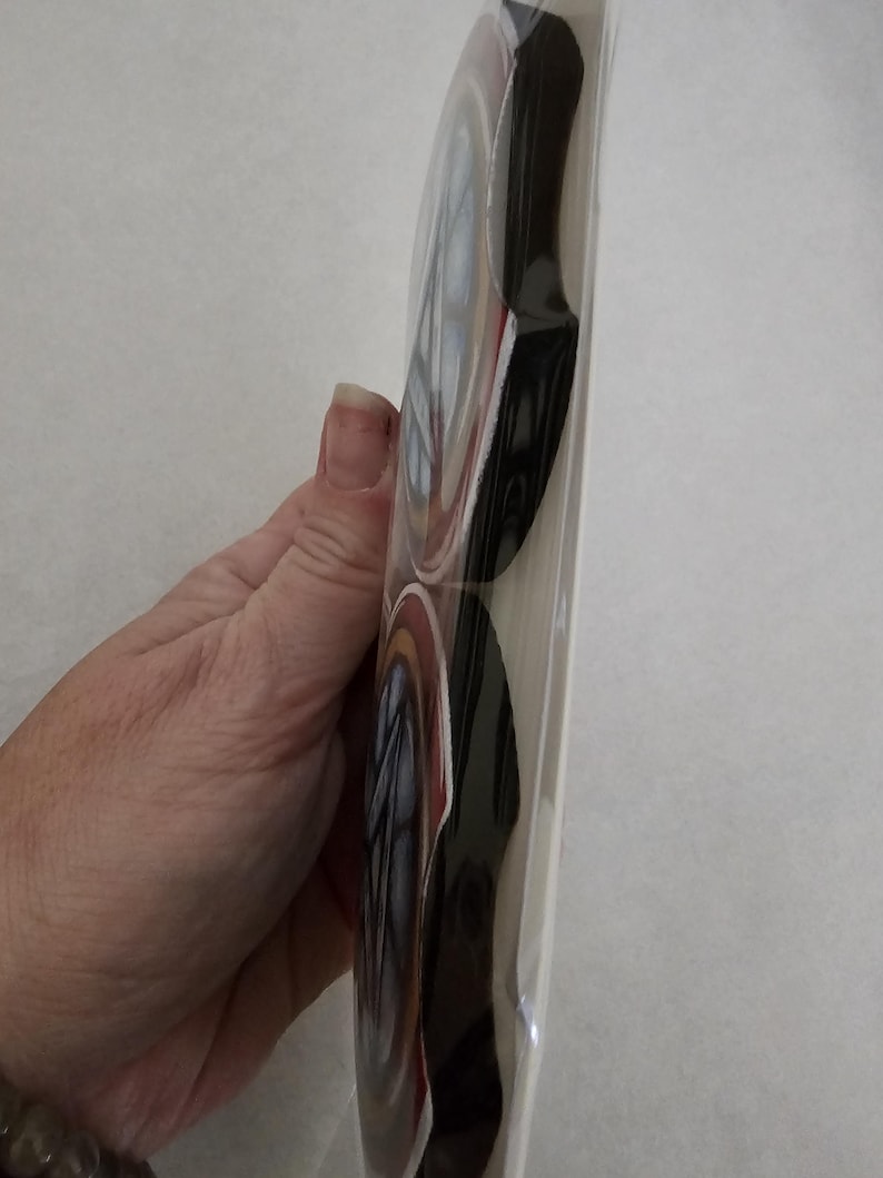 May include: A stack of round, decorative coasters in a clear plastic sleeve. The coasters have a design with red, white, and blue accents, and a black border. The coasters are held by a person's hand.