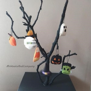 May include: A black branch tree with felt Halloween ornaments hanging from it. The ornaments include a ghost, a pumpkin, a witch's hat, a candy corn, a Frankenstein, and a spaceship. The tree is on a black square base. MrHootsAndCompany is written on the wall.