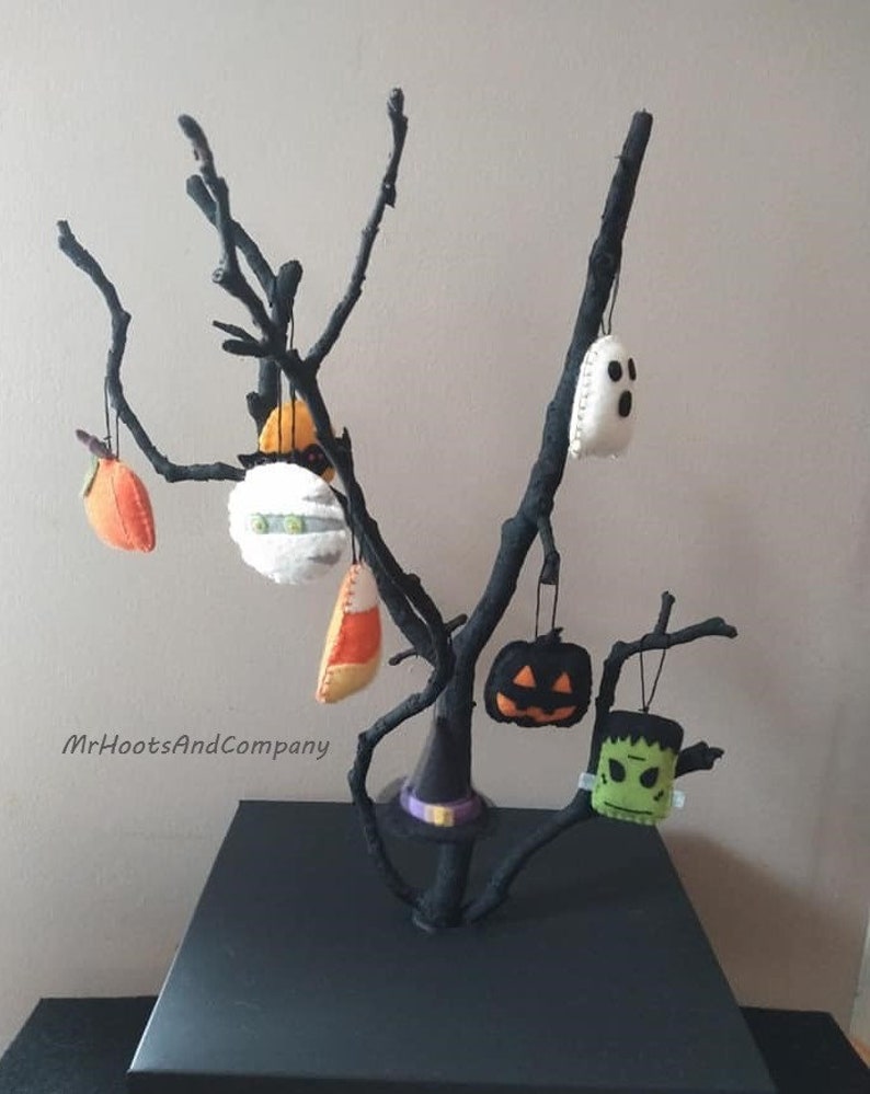 May include: A black branch tree with felt Halloween ornaments hanging from it. The ornaments include a ghost, a pumpkin, a witch's hat, a candy corn, a Frankenstein, and a spaceship. The tree is on a black square base. MrHootsAndCompany is written on the wall.