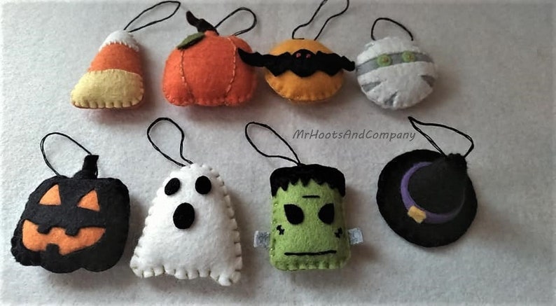 May include: Nine felt Halloween ornaments with hanging loops. The ornaments are shaped like a pumpkin, ghost, witch's hat, candy corn, Frankenstein, mummy, bat, and two pumpkins.  MrHootsAndCompany is printed on the white background.