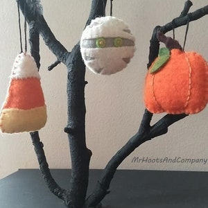 May include: Three felt Halloween ornaments hanging from a black branch. The ornaments are a mummy, a pumpkin, and a candy corn. The ornaments are handmade and have a rustic, vintage look.