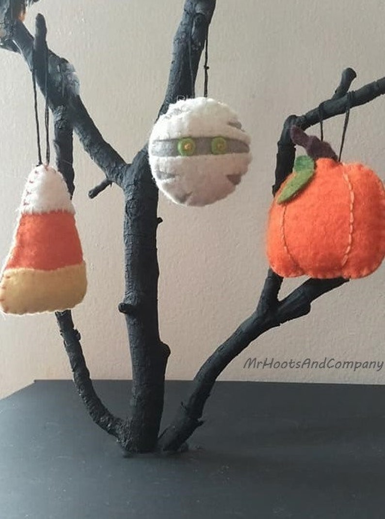 May include: Three felt Halloween ornaments hanging from a black branch. The ornaments are a mummy, a pumpkin, and a candy corn. The ornaments are handmade and have a rustic, vintage look.