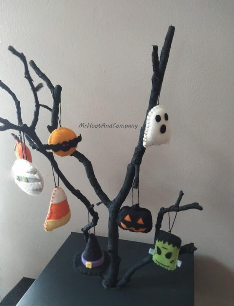 May include: A set of felt Halloween ornaments hanging from a black branch. The ornaments include a ghost, a pumpkin, a witch's hat, a candy corn, a Frankenstein, and a vampire.