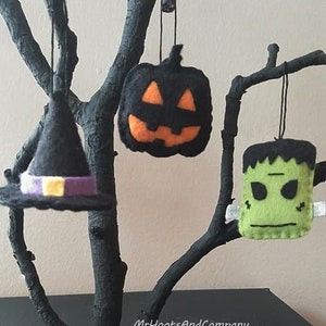 May include: Three felt Halloween ornaments hanging from a black branch. The ornaments are a witch's hat with a purple band, a jack-o'-lantern with an orange face, and a green Frankenstein's monster.