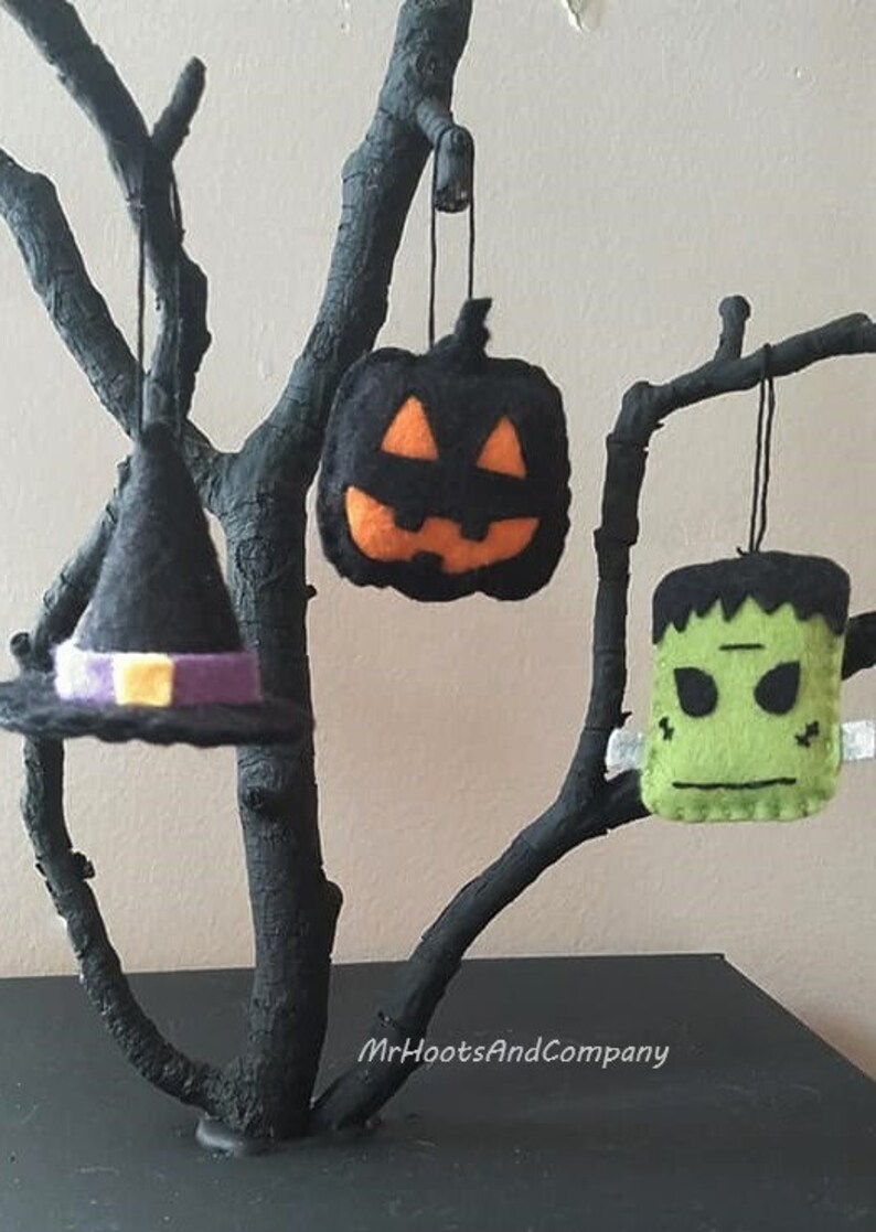 May include: Three felt Halloween ornaments hanging from a black branch. The ornaments are a witch's hat with a purple band, a jack-o'-lantern with an orange face, and a green Frankenstein's monster.