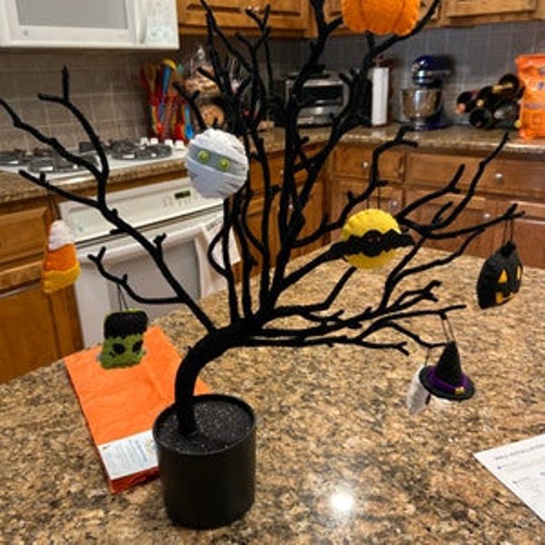 May include: A black twig tree with Halloween-themed felt ornaments. The ornaments include a mummy, a pumpkin, a witch's hat, a candy corn, a Frankenstein, and a black cat.