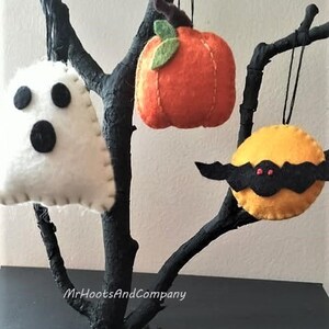 May include: Three felt Halloween ornaments hanging from a black branch. The ornaments are a white ghost with black eyes and mouth, an orange pumpkin with a green stem, and a yellow bat with black wings and red eyes.