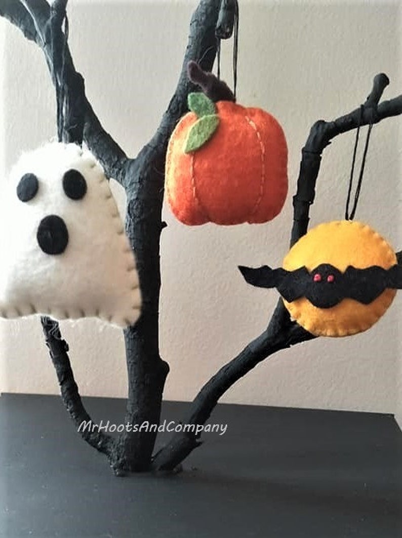 May include: Three felt Halloween ornaments hanging from a black branch. The ornaments are a white ghost with black eyes and mouth, an orange pumpkin with a green stem, and a yellow bat with black wings and red eyes.