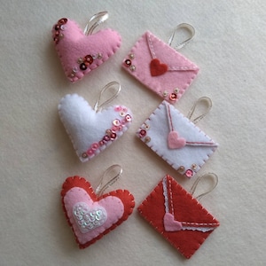 May include: A collection of handmade felt ornaments in the shape of hearts and envelopes. The hearts are pink, white, and red, embellished with sequins. The envelopes are pink and white, with red heart accents and silver ribbon loops.