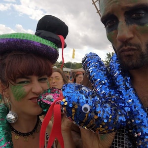 Peut inclure: Une personne porte un serpent à paillettes avec un chapeau haut de forme noir et une langue rouge. Une autre personne tient un serpent à paillettes bleu. Les deux ont du maquillage. Le fond montre un ciel nuageux et d'autres personnes.