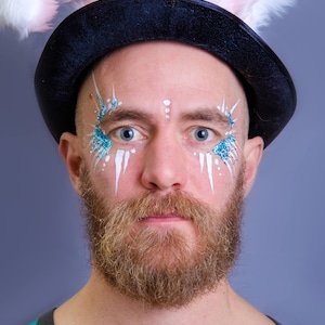 May include: A man wearing a black top hat with pink bunny ears, white and blue face paint, and a brown beard.