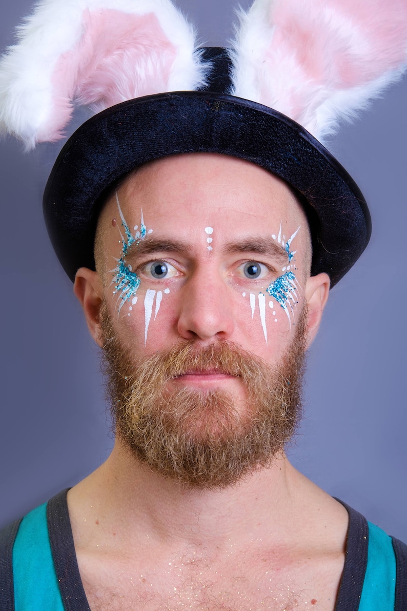 May include: A man wearing a black top hat with pink bunny ears, white and blue face paint, and a brown beard.