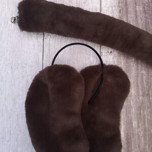 Op de afbeelding: Een bruine faux fur staart met een zilveren sluiting en bijpassende oorbeschermers. De oorbeschermers hebben een zwarte hoofdband. De set is gemaakt van zacht, pluche materiaal.