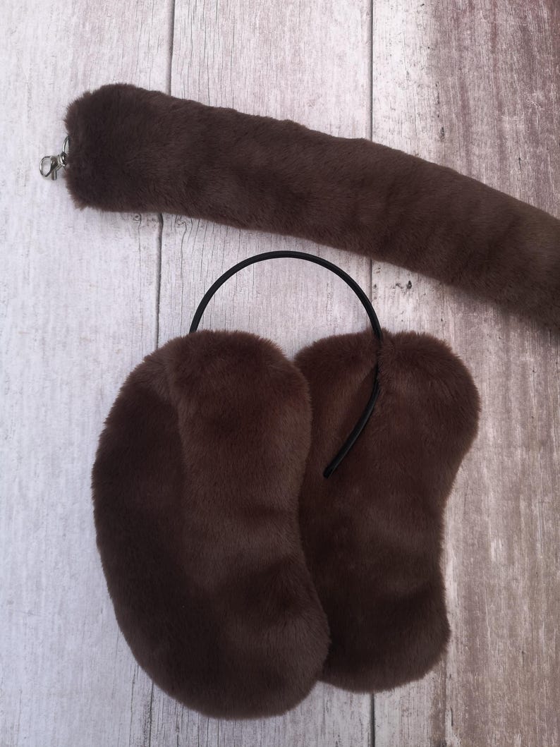 Op de afbeelding: Een bruine faux fur staart met een zilveren sluiting en bijpassende oorbeschermers. De oorbeschermers hebben een zwarte hoofdband. De set is gemaakt van zacht, pluche materiaal.