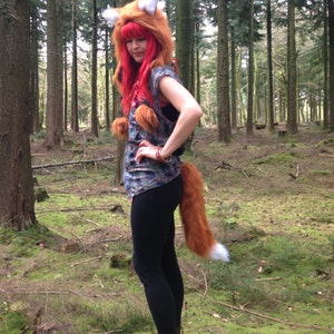 May include: A woman wearing a fox costume with a bushy tail and a red wig in a forest setting.