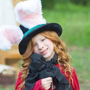 May include: A young girl wearing a red jacket and a black bow tie, with a black top hat with white bunny ears. The hat has a teal band around the base.
