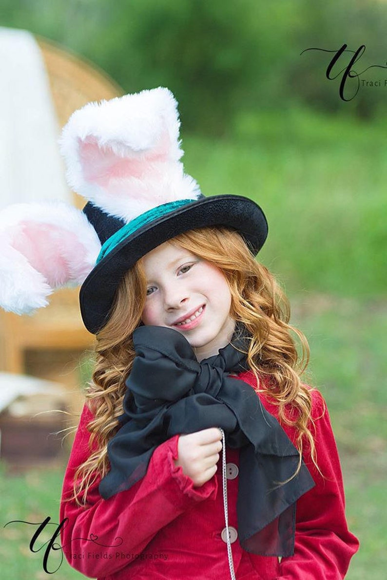 May include: A young girl wearing a red jacket and a black bow tie, with a black top hat with white bunny ears. The hat has a teal band around the base.