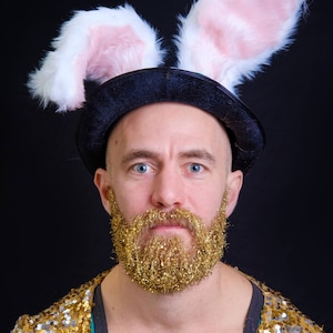 May include: A man wearing a black top hat with pink bunny ears, a gold glitter beard, and a gold sequined jacket.