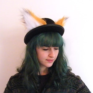 May include: A person with green hair wearing a black hat with faux fur fox ears attached. The hat has a wide brim and the fur is white, orange, and black.