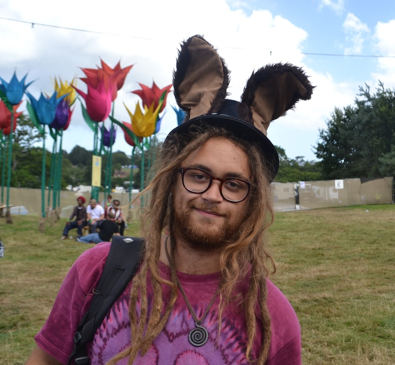 May include: A person with long brown dreadlocks wearing a pink and purple tie-dye shirt and a black top hat with brown bunny ears. The person is wearing round glasses and has a beard.