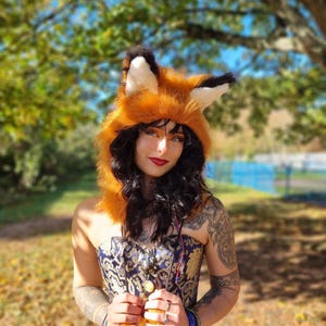 May include: A plush orange and white faux fur hood featuring pointed fox ears with black tips. The hood is designed to frame the head with soft, textured material, mimicking the appearance of a fox, and includes long side panels.