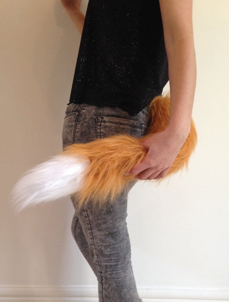 May include: A fluffy orange and white fox tail costume accessory. The tail is made of soft, plush fur and has a white tip. It is attached to a person's waist with a belt.