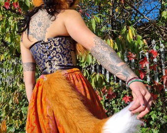 May include: A faux fur fox hat with pointed ears and a matching long, bushy orange tail with a white tip. The ensemble is paired with a blue and gold brocade corset featuring black lace-up detailing and a tiered orange and pink patterned skirt.