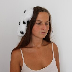 Op de afbeelding: Een witte haarband met zwarte vlekken, gemaakt van zacht, pluizig materiaal. Het accessoire heeft een Dalmatische patroon. Deze speelse haarband is een leuk mode-item, geschikt voor verschillende gelegenheden.