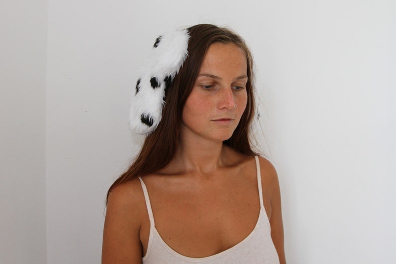 Op de afbeelding: Een witte haarband met zwarte vlekken, gemaakt van zacht, pluizig materiaal. Het accessoire heeft een Dalmatische patroon. Deze speelse haarband is een leuk mode-item, geschikt voor verschillende gelegenheden.