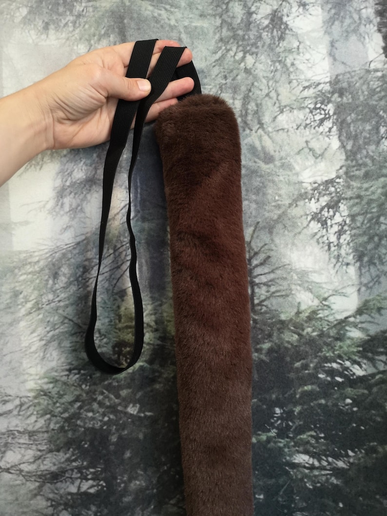 May include: A brown faux fur tail with a black adjustable strap. The tail is long and fluffy, and the strap is made of a durable material.