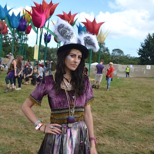 May include: A woman wearing a purple velvet top with gold trim and a silver skirt. She is wearing a white bunny ear headband and has glitter makeup on her face. She is standing in a field with large colorful flower sculptures in the background.