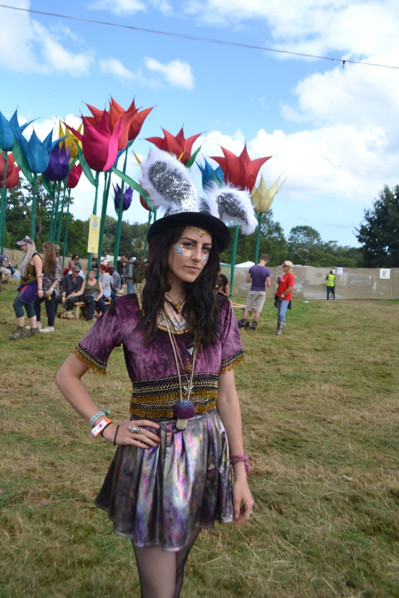 May include: A woman wearing a purple velvet top with gold trim and a silver skirt. She is wearing a white bunny ear headband and has glitter makeup on her face. She is standing in a field with large colorful flower sculptures in the background.
