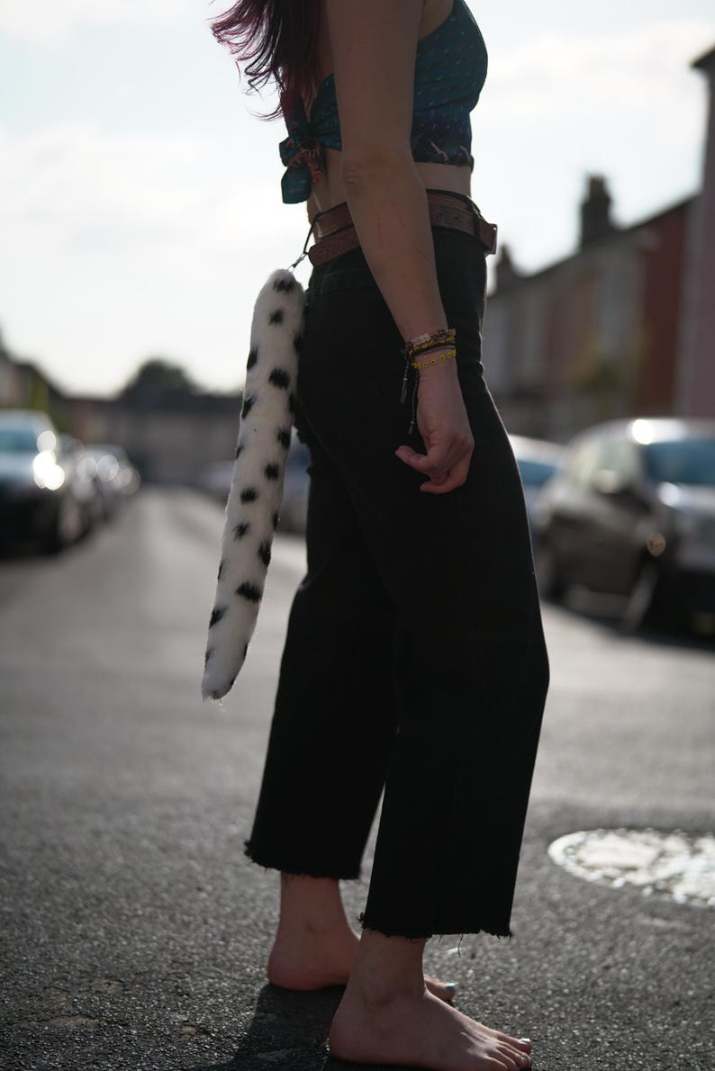 May include: A person wearing black pants and a cropped top with a white and black spotted tail attached to their belt.