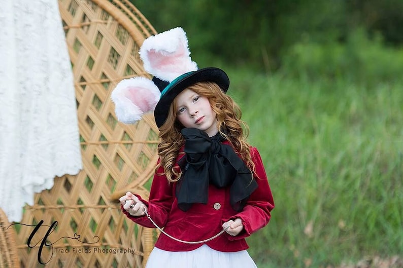 May include: A young girl dressed as the Mad Hatter from Alice in Wonderland. She is wearing a red velvet jacket, a black bow tie, and a black top hat with white bunny ears. She is holding a pocket watch on a chain.