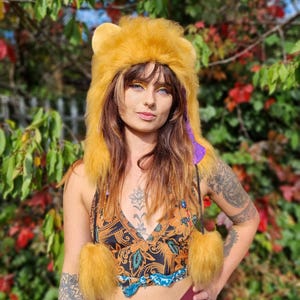 May include: A golden yellow faux fur hood featuring rounded ears and two long side tassels with matching pom-poms. The hood is lined with a purple patterned fabric and includes thin black braided ties with small colorful beads attached.