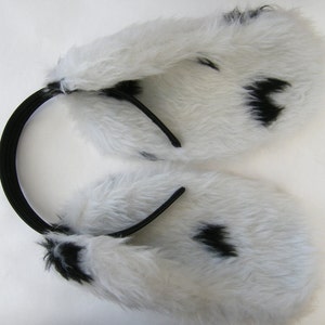 Op de afbeelding: Witte imitatiebont oorbeschermers met zwarte vlekken, bevestigd aan een zwarte hoofdband. De oorbeschermers zijn zacht en pluizig, met een patroon dat lijkt op een Dalmatische hond. De hoofdband is gemaakt van flexibel, elastisch materiaal.