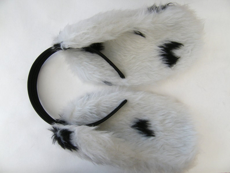 Op de afbeelding: Witte imitatiebont oorbeschermers met zwarte vlekken, bevestigd aan een zwarte hoofdband. De oorbeschermers zijn zacht en pluizig, met een patroon dat lijkt op een Dalmatische hond. De hoofdband is gemaakt van flexibel, elastisch materiaal.