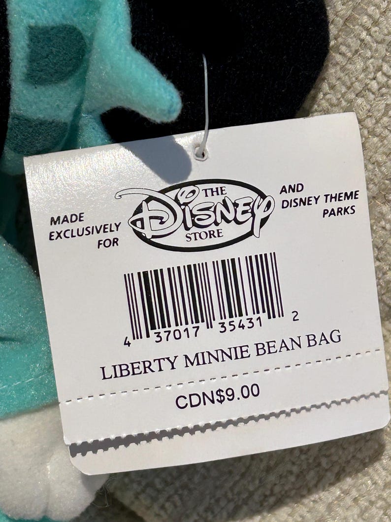 May include: A white tag with black text and a barcode. The tag says "Made Exclusively For The Disney Store" and "And Disney Theme Parks". The tag also says "Liberty Minnie Bean Bag" and "CDN$9.00".